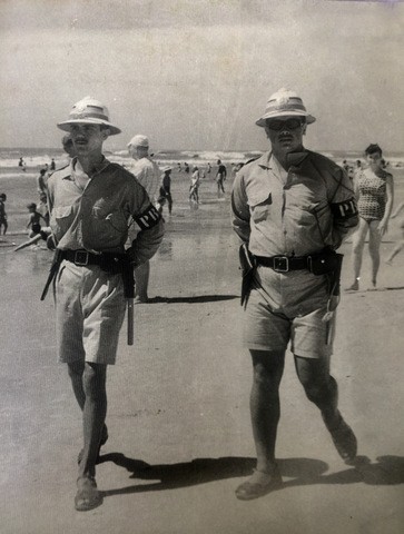 Dois oficiais uniformizados, vestindo shorts e chapéu claro com abas, na praia e grupo de pessoas ao fundo. 
