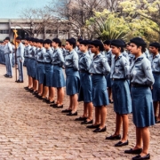 Grupo de alunos do colégio Tiradentes, separados por gênero feminino e masculino, posicionados em fila lateral, uniformizados, no pátio do colégio.