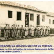 grupo de oficiais da Brigada uniformizados com calça e chapéu, posicionados lado a lado, em frente a uma casa.