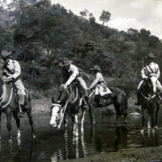 Grupo de homens vestindo uniforme e chapéu com abas, atravessando um lago a cavalo.