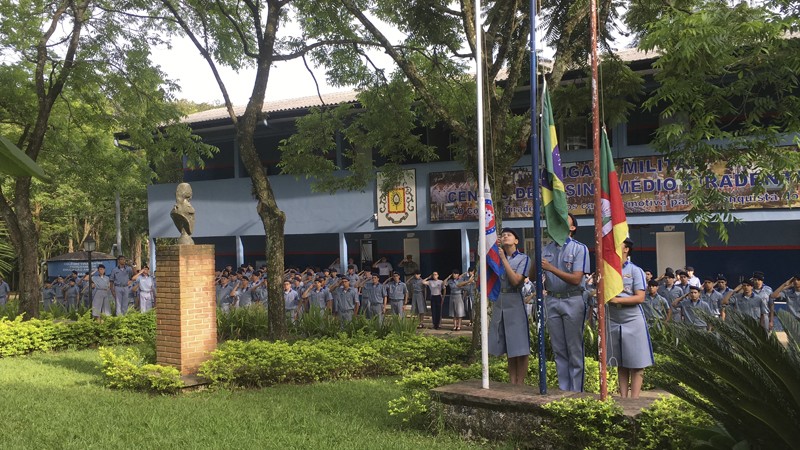 Foto da fachada do prédio do colégio, com os alunos em forma, e outros astiando a bandeira