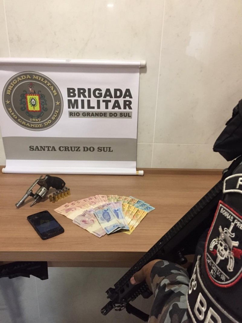 Rev&oacute;lver, celular e c&eacute;dulas de dinheiro no fundo um banner da Brigada Militar
