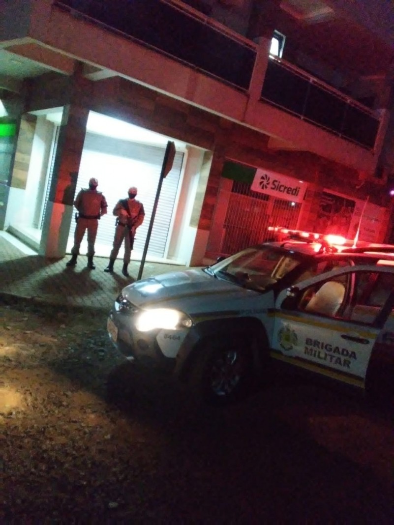 Uma viatura da Brigada Militar a frente e dois policiais militares ao fundo fazendo policiamento ostensivo em frente a uma ag&ecirc;ncia banc&aacute;ria