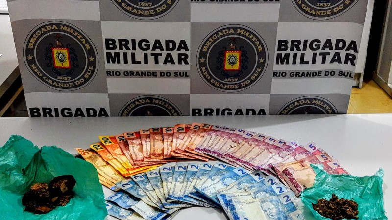 Sobre uma mesa branca drogas e dinheiro em esp&eacute;cie. No fundo o banner da Brigada Militar.
