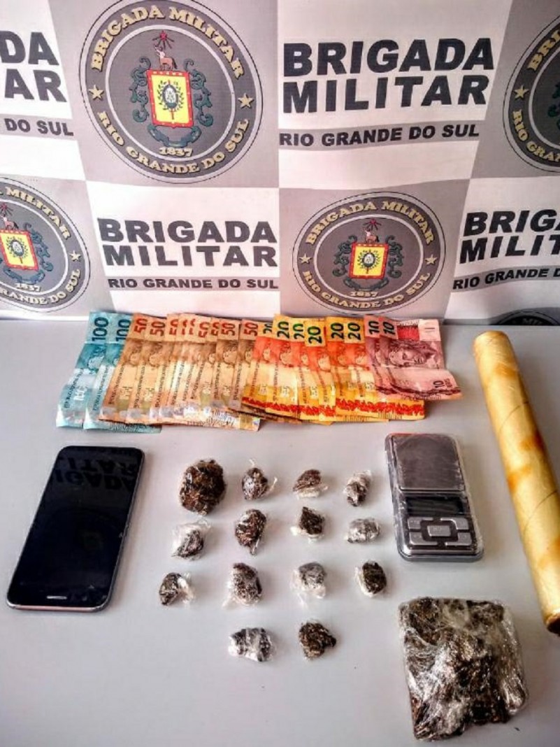 Sobre uma mesa branca esta celular, balan&ccedil;a de precis&atilde;o, c&eacute;dulas de dinheiro e drogas apreendidas. Ao fundo o banner da brigada Militar.