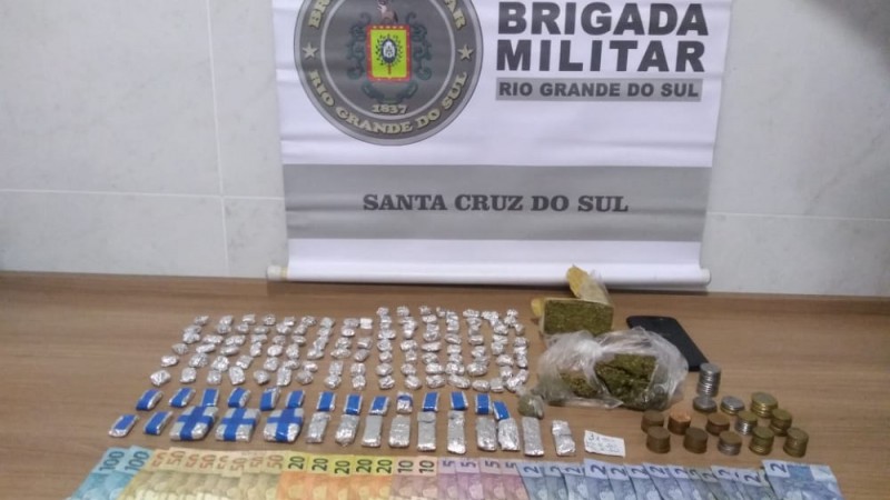 Sobre uma mesa drogas fracionadas, celular e dinheiro em esp&eacute;cie e no fundo o banner da Brigada Militar