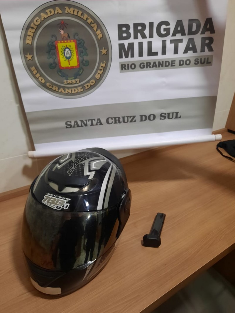 Sobre uma mesa um capacete preto e um carregador de pistola. No fundo o banner da Brigada Militar.