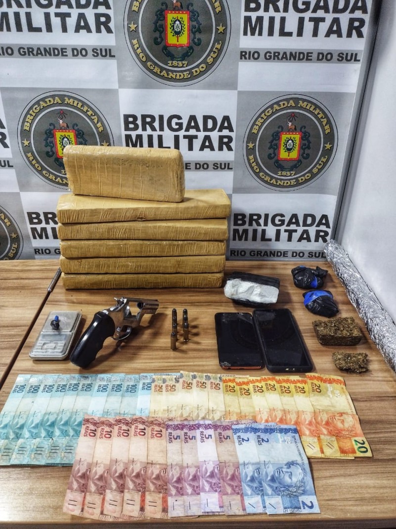 Sobre uma mesa tijolos de drogas e por&ccedil;&otilde;es fracionadas. Moeda em especie, celulares, uma arma de fogo e muni&ccedil;&otilde;es ao lado. No fundo o banner institucional da Brigada Militar.