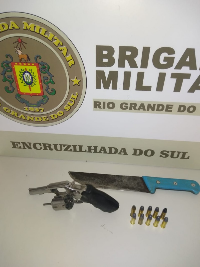 Ao fundo da imagem o banner da Brigada Militar. Sobre uma mesa uma arma de fogo, muni&ccedil;&otilde;es e uma faca.