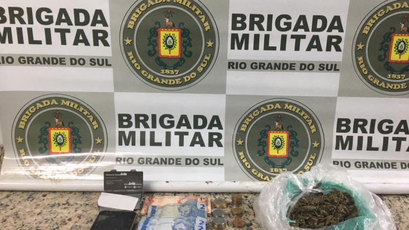 Ao fundo o banner da Brigada Militar. Sobre uma mesa drogas, moeda em esp&eacute;cie e um celular.
