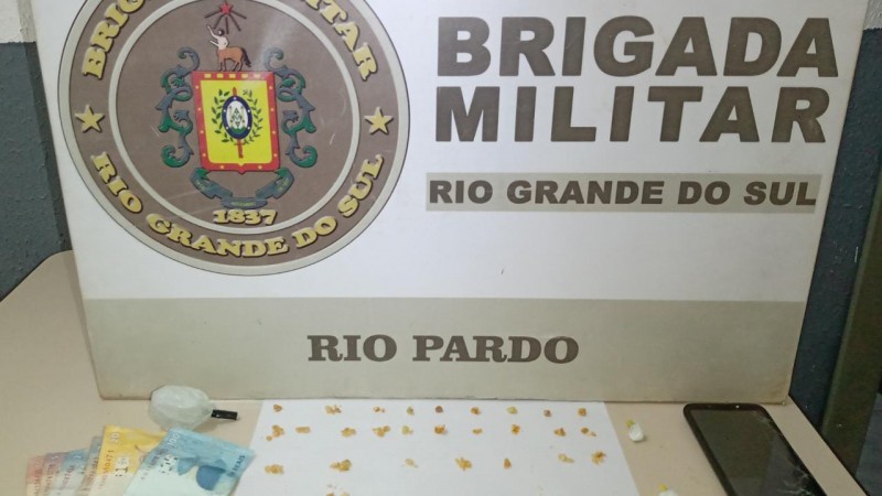 No fundo o banner da Brigada Militar. Sobre uma mesa drogas, celular e moeda em esp&eacute;cie.