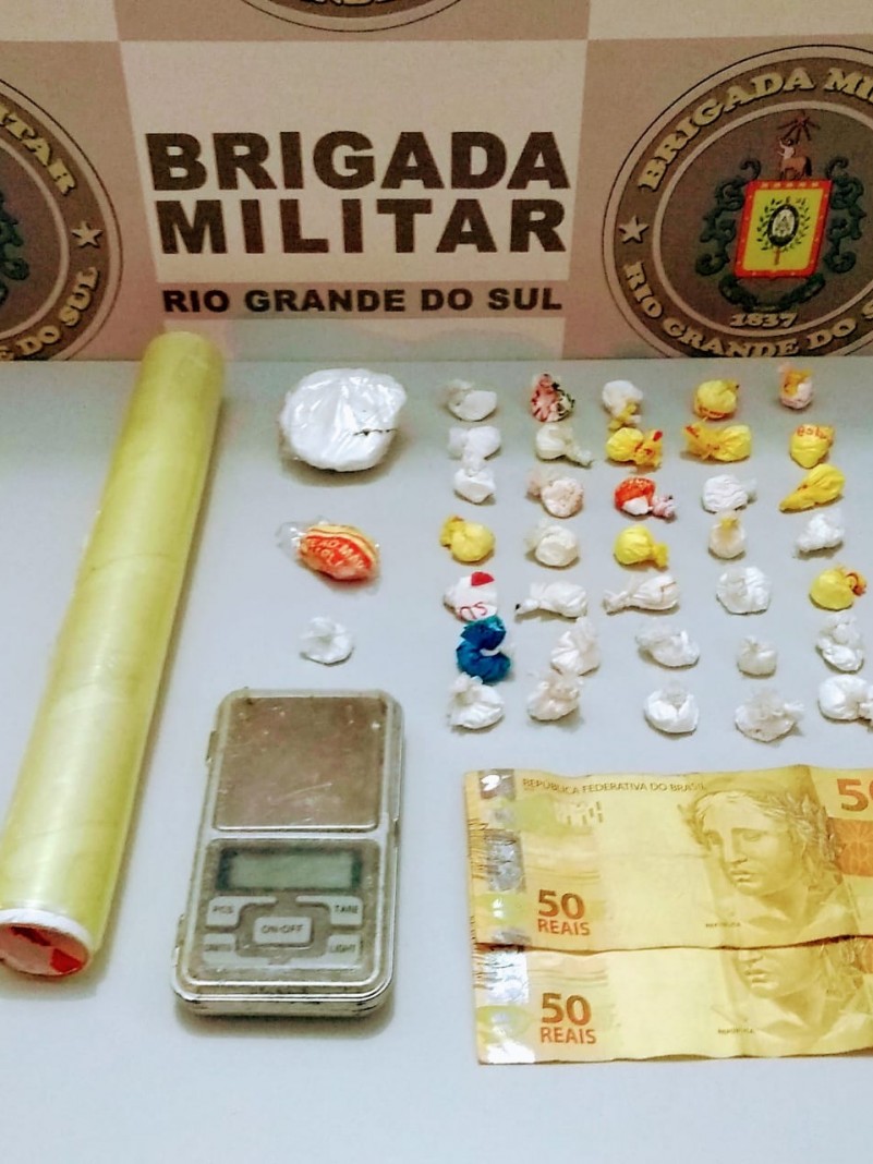 No fundo o banner da Brigada Militar. Sobre uma mesa drogas fracionadas, moeda em esp&eacute;cie e uma balan&ccedil;a de precis&atilde;o.