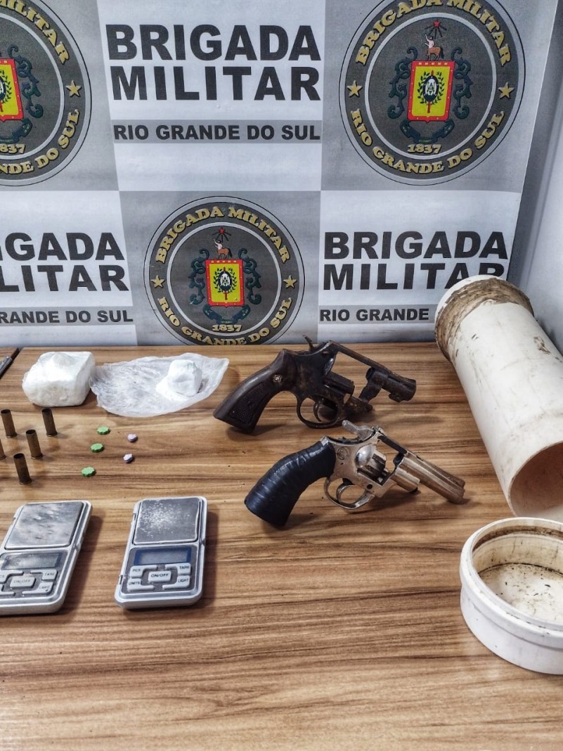 No fundo o banner da Brigada Militar. Sobre uma mesa armas, balan&ccedil;a de precis&atilde;o, drogas, cano de PVC e cartuchos de muni&ccedil;&atilde;o.