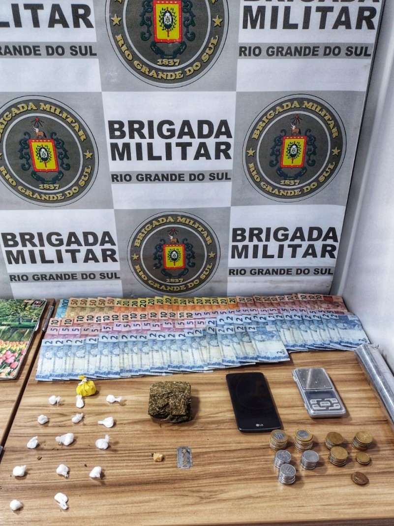 Ao fundo o banner da Brigada Militar. Sobre uma mesa drogas fracionadas, celulares, dinheiro em esp&eacute;cie, balan&ccedil;a de precis&atilde;o.