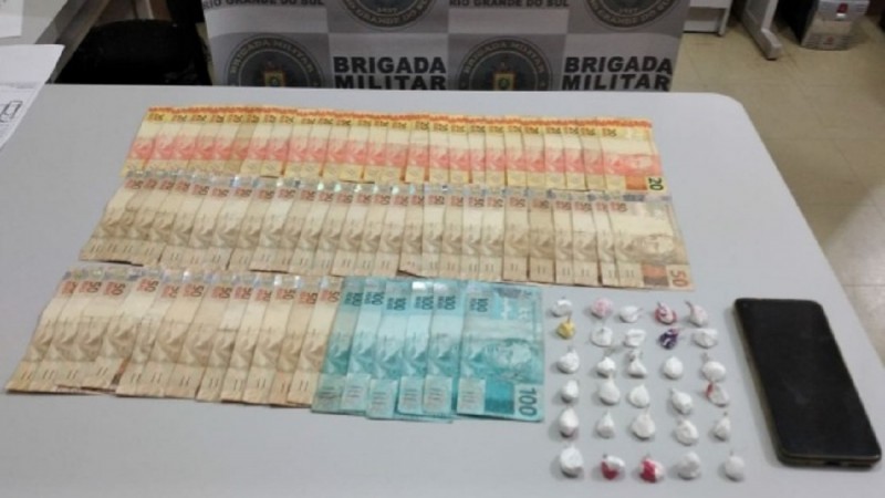 Ao fundo o banner da Brigada Militar. Sobre uma mesa dinheiro em esp&eacute;cie, drogas fracionadas e um celular.