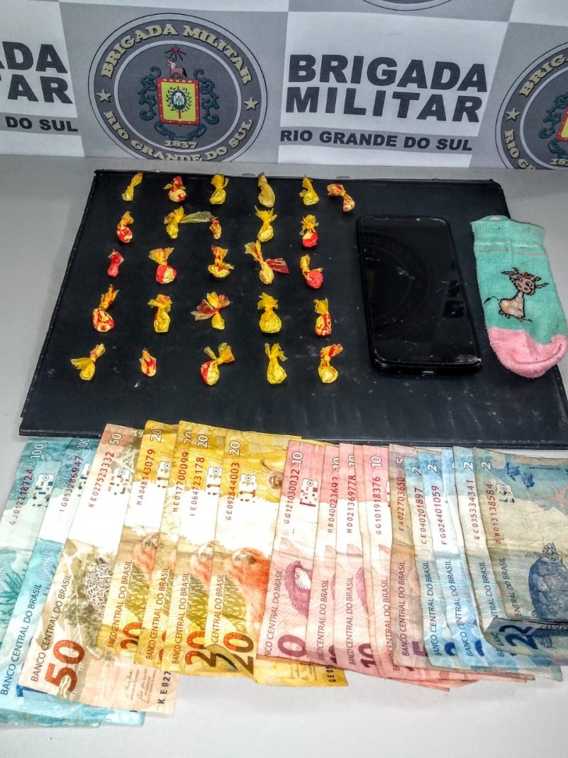 Ao fundo o banner da Brigada Militar. Sobre uma mesa drogas fracionadas, dinheiro em esp&eacute;cie e celular. 