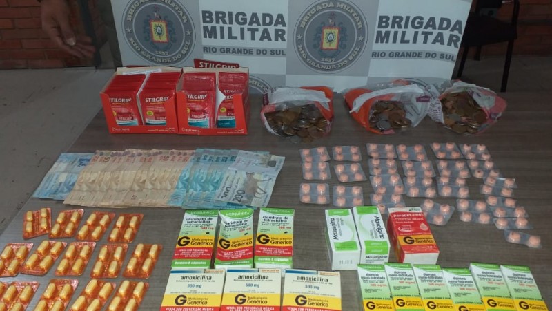 Ao fundo o banner da Brigada Militar. Sobre uma mesa diversas caixas de rem&eacute;dios  e comprimidos avulsos, bem como moeda em esp&eacute;cie