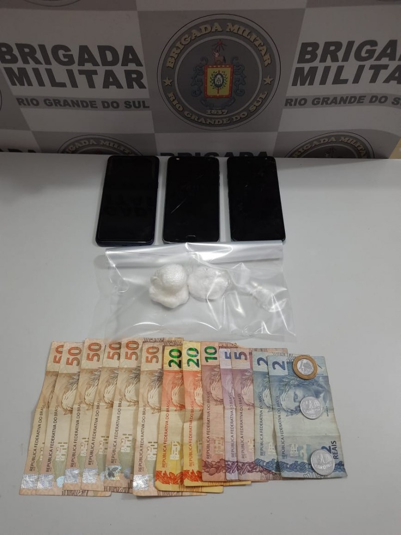 Ao fundo o banner da Brigada Militar. Sobre uma mesa tr&ecirc;s celulares, drogas fracionadas e dinheiro em esp&eacute;cie.
