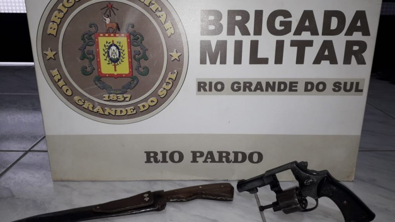 Ao fundo o banner da Brigada Militar. Em sua frente uma faca com bainha e uma arma de fogo.