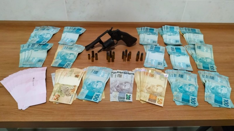 Sobre uma mesa dinheiro em esp&eacute;cie, uma arma de fogo e muni&ccedil;&otilde;es ao lado.