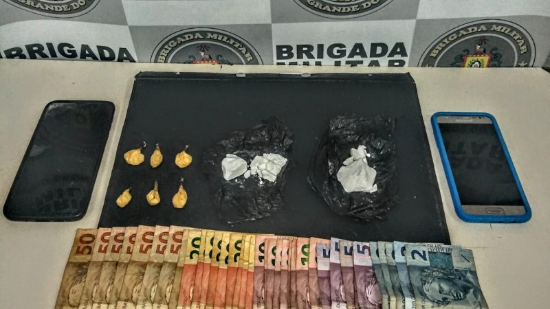 Ao fundo o banner da Brigada Militar, e sobre uma mesa drogas fracionadas, dois celulares e dinheiro em esp&eacute;cie.