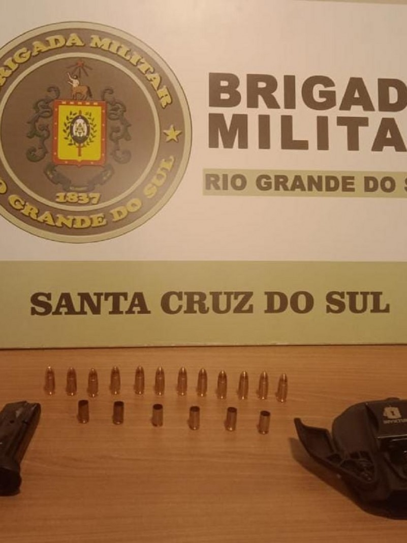 Doze muni&ccedil;&otilde;es intactas e a frente seis estojos de muni&ccedil;&otilde;es deflagradas. Ao lado um carregador e no outro lado um porta carregador. Ao fundo o banner institucional da Brigada Militar de Santa Cruz do Sul.