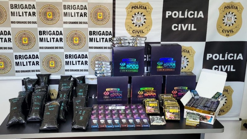 Na imagem sob uma mesa está o material apreendido, dentre drogas, 43 kg, embalagens que eram utilizadas para esconder os ilícitos. Ao fundo, na parede os banners da Brigada Militar e polícia civil.