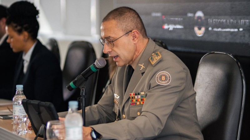 Foto mostra o comandante-geral da BM, coronel Cláudio dos Santos Feoli, sentado em uma mesa, em frente a um microfone, lendo um discurso