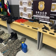 Foto mostra três mesas com munições e armas de fogo. No chão, na frente da mesa, há uniformes militares.