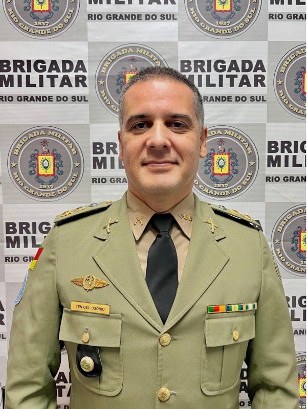 Na imagem, o comandante do 20° BPM está posicionado em frente ao banner da Brigada Militar e vestido com o traje formal da Brigada Militar, na cor bege. O fardamento possui medalhas e detalhes em dourado nos ombros, que representam o posto de Tenente-coronel. 