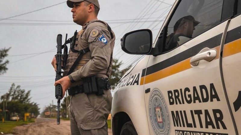 Foto mostra um policial militar de perfil segurando um fuzil ao lado de uma viatura