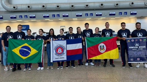 Foto mostra os alunos enfileirados no embarque do aeroporto, segurando as bandeiras do Brasil, do Colégio Tiradentes e do Rio Grande do Sul