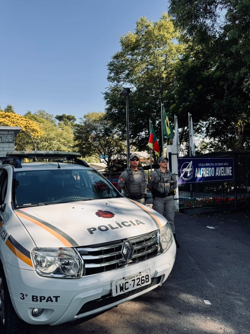 Foto mostra dupla de policiais militares ao lado de uma viatura em frente a uma escola municipal.