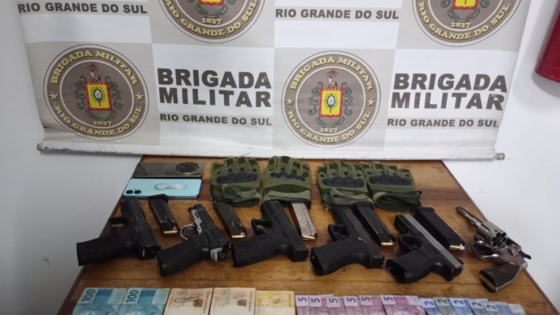Mesa com os materiais apreendidos pela polícia: um revólver, cinco pistolas, dinheiro, luvas, dois telefones celulares. Mostra um banner com o brasão da Brigada Militar