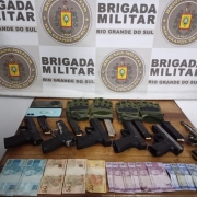 Mesa com os materiais apreendidos pela polícia: um revólver, cinco pistolas, dinheiro, luvas, dois telefones celulares. Mostra um banner com o brasão da Brigada Militar