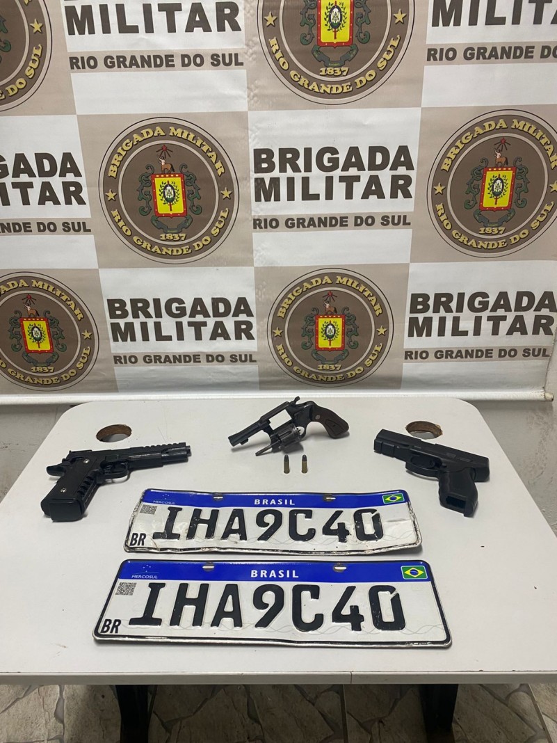 Apreensões sobre uma mesa: três armas, munições e duas placas de veículos. Ao fundo está o brasão da Brigada Militar