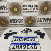 Apreensões sobre uma mesa: três armas, munições e duas placas de veículos. Ao fundo está o brasão da Brigada Militar