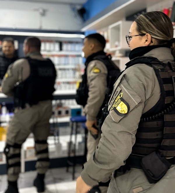 Na imagem constam três policiais militares atuando na fiscalização de uma loja de celulares.