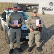 Policiais Militares masculino e feminino lado a lado, mostram o folder informativo que distribuíram aos motoristas