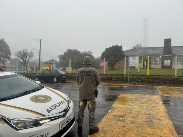 Foto mostra policial em frente a uma escola, ao lado de uma viatura, durante um dia de chuva