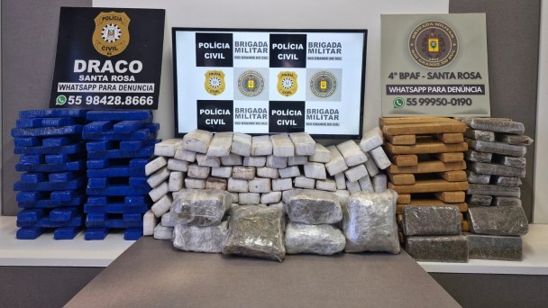 Tabletes de maconha nas cores azul, branco, amarelo e transparente sobre uma mesa. Ao fundo, aparecem os banners da Draco, Polícia Civil e BM de Santa Rosa