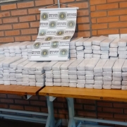 Tabletes de maconha embalados nas cores branca e azul, empilhados e dispostos sobre uma mesa, com o banner da BM pendurado na parede 