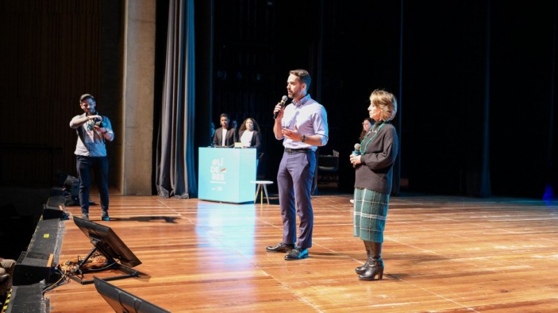 No palco com luzes refletidas, aparecem o governador e a secretária em pé, falando ao microfone