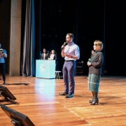 No palco com luzes refletidas, aparecem o governador e a secretária em pé, falando ao microfone