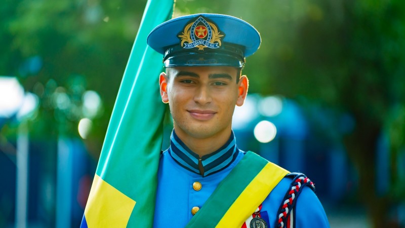 Jovem estudante, sorridente, aparece fardado e carregando a bandeira brasileira