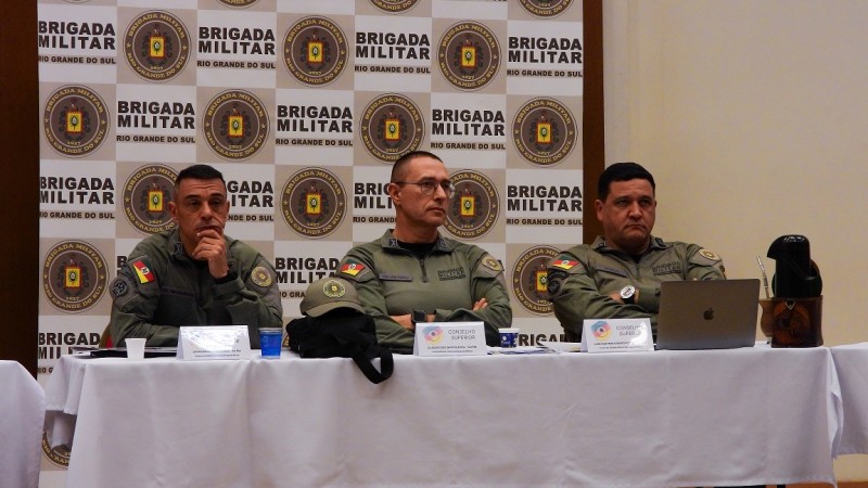 os três comandantes estão sentados, assistindo alguém falar, há uma mesa com toalha branca e ao fundo está um banner com o brasão da BM