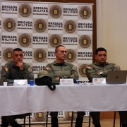 os três comandantes estão sentados, assistindo alguém falar, há uma mesa com toalha branca e ao fundo está um banner com o brasão da BM
