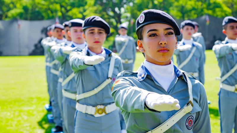 alunas estão em posição, em filas, uniformizadas, em um gramado, numa cerimônia de formatura