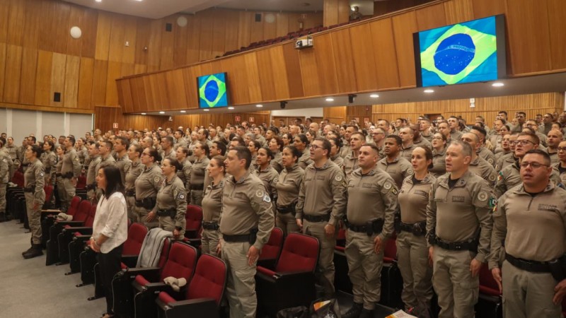 Policiais militares que atuam no Proerd, compondo o público do evento, durante execução do Hino Nacional, na abertura do evento