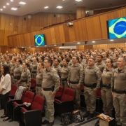 Policiais militares que atuam no Proerd, compondo o público do evento, durante execução do Hino Nacional, na abertura do evento